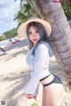 A woman in a hat leaning against a palm tree on the beach.